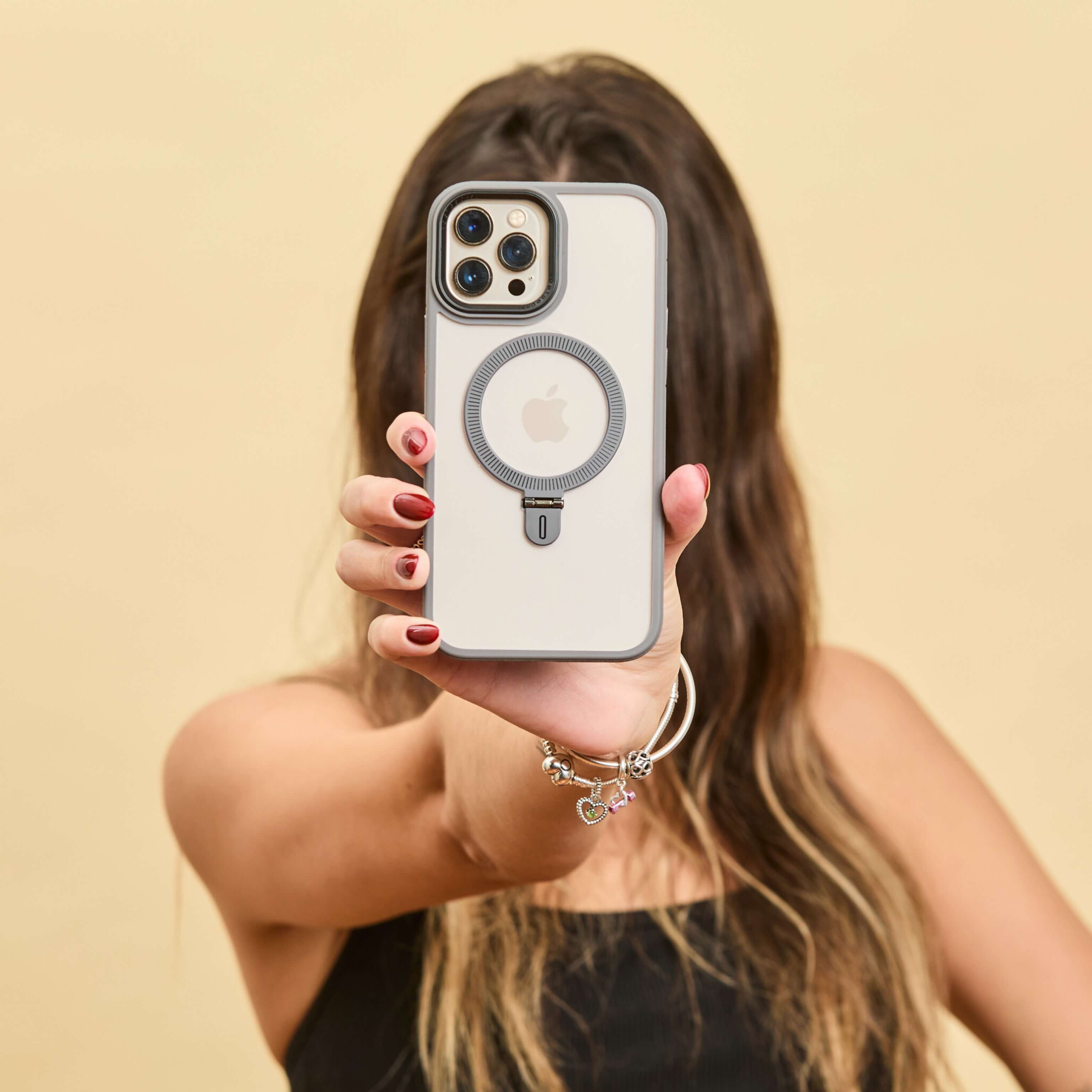 Funda gris con anillo trasero sostenida de frente por una mujer, rostro cubierto por el teléfono, fondo beige. #color_gris