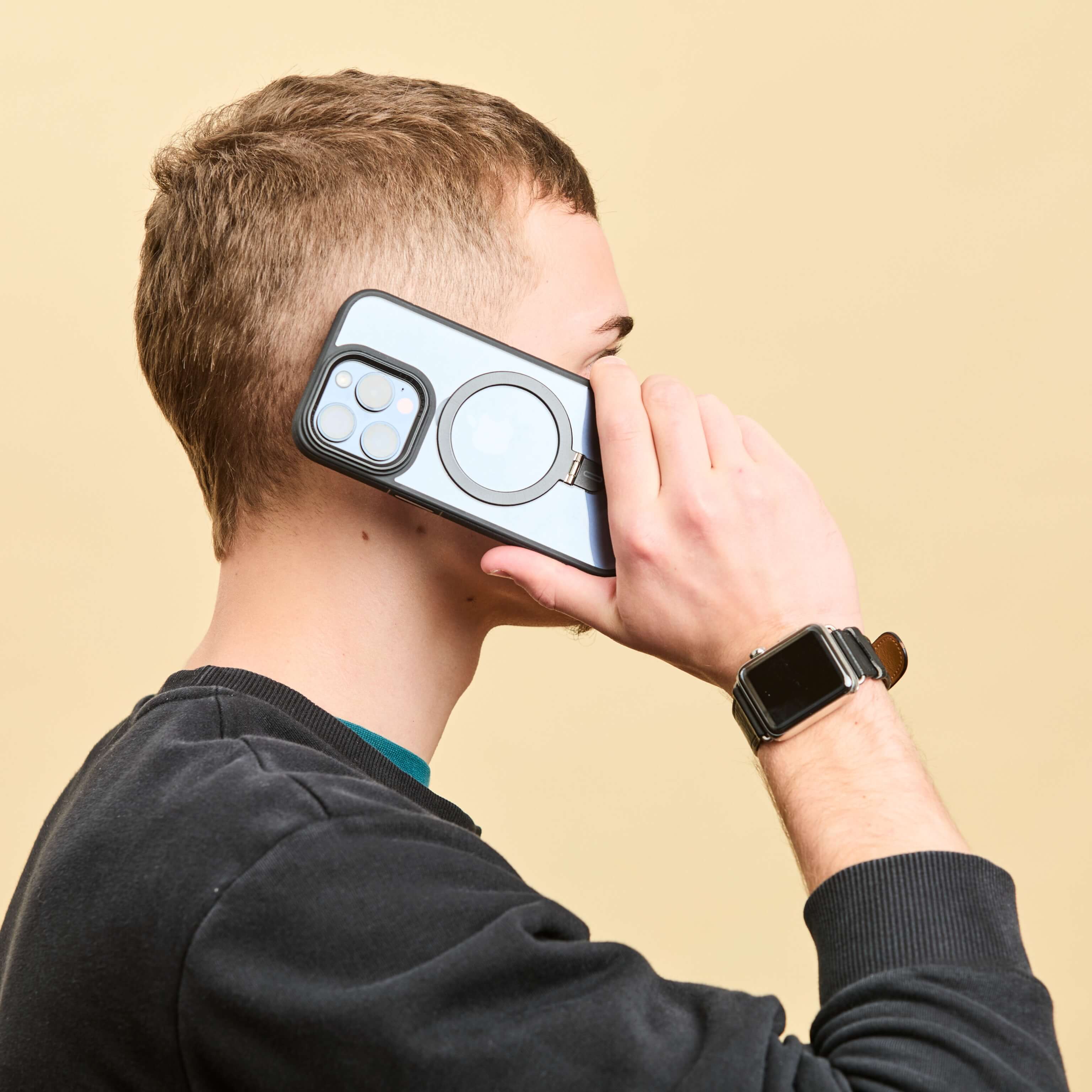 Hombre sosteniendo un iPhone con funda gris y anillo trasero junto a su oreja, simulando una llamada, sobre fondo beige. #color_negro