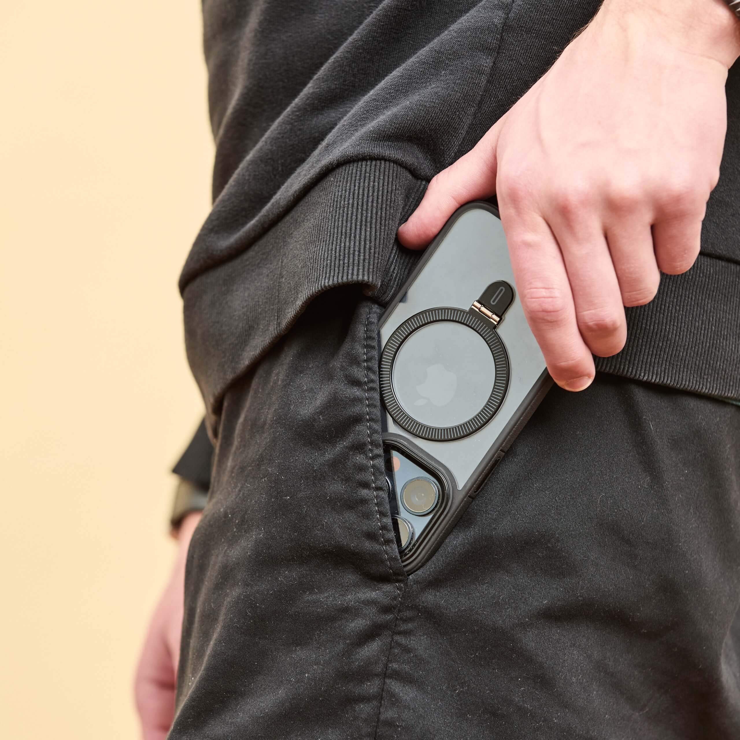 Funda negra con anillo en un iPhone parcialmente guardado en el bolsillo de un pantalón negro, sostenido por una mano masculina. #color_negro