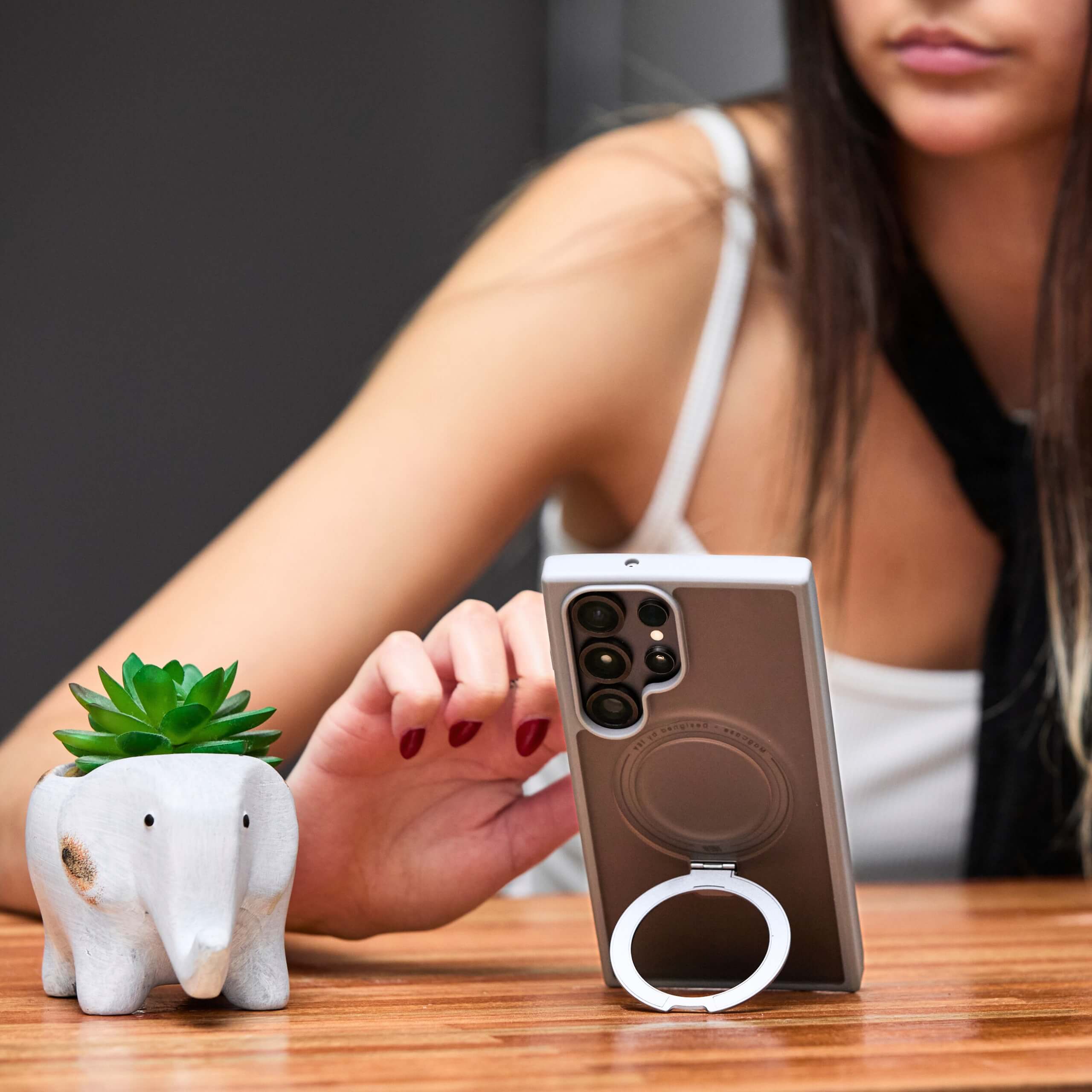 Mujer usando el teléfono sobre la mesa con funda para Samsung S25 Ultra y aro magnético, apoyada con el aro como soporte. #color_gris