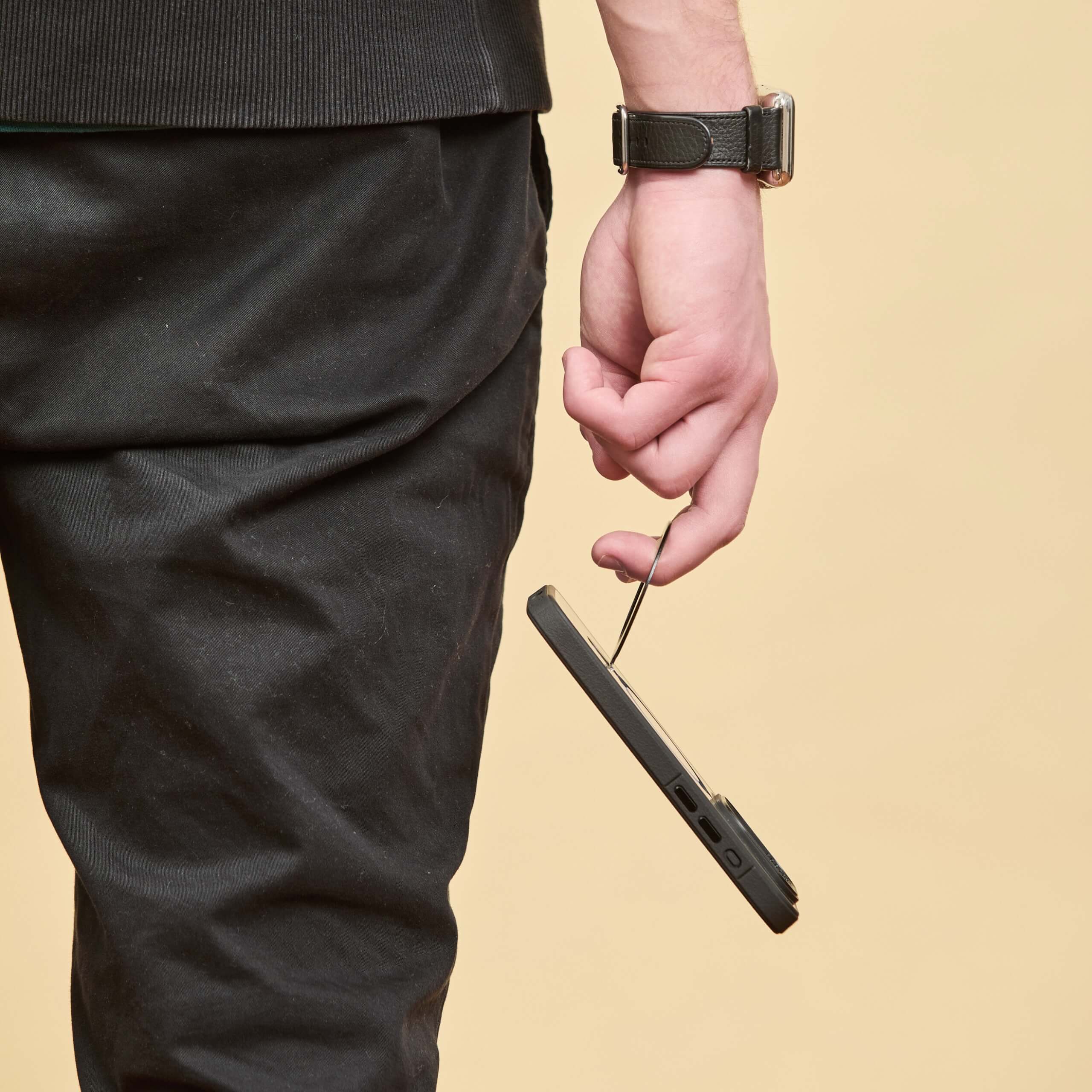 Funda negra colgando desde la mano de un hombre, sostenida por el anillo trasero, con pantalón oscuro y fondo beige. #color_negro