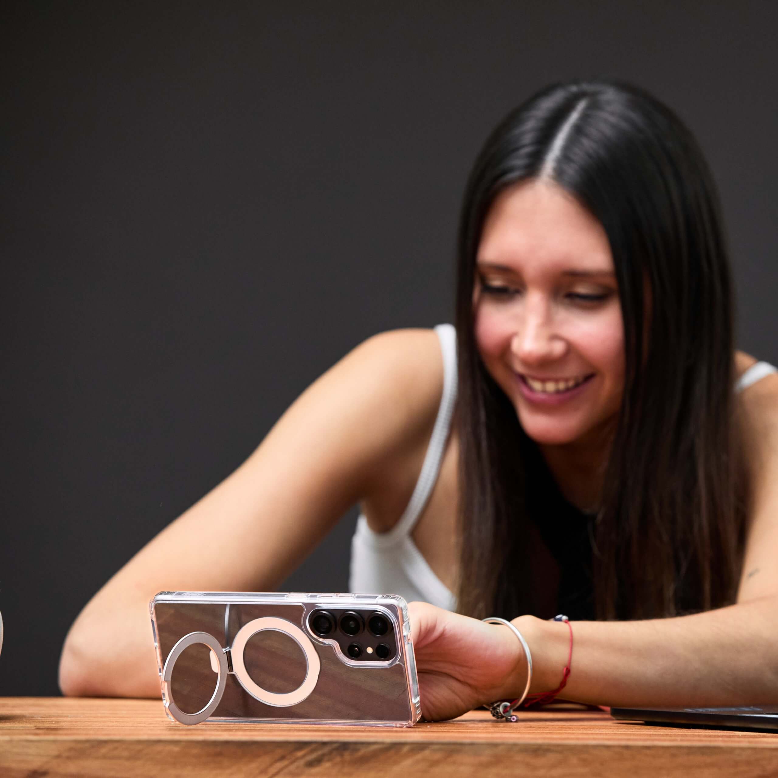 Mujer apoyada en la mesa mirando el teléfono con funda MagSafe para Samsung, con expresión sonriente. #color_transparente