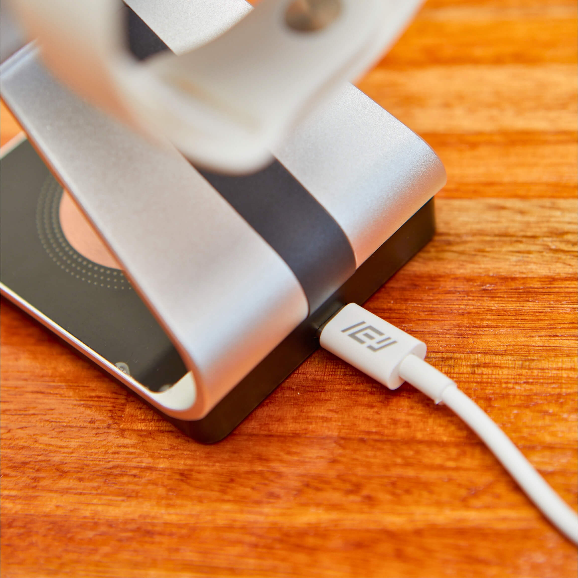 Cable USB-C IEY blanco conectado a base de carga metálica para smartphone, sobre superficie de madera - soporte moderno para escritorio