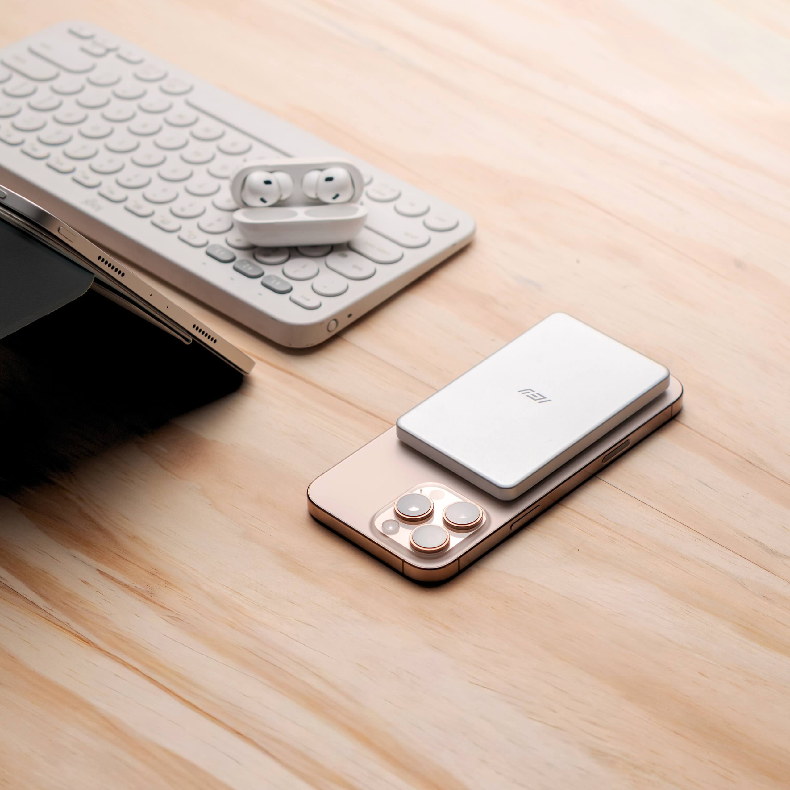 Batería inalámbrica ultrafina IEY MagPower en color blanco sobre iPhone, acompañada de teclado y auriculares, estilo lifestyle en escritorio de madera. #color_gris plateado