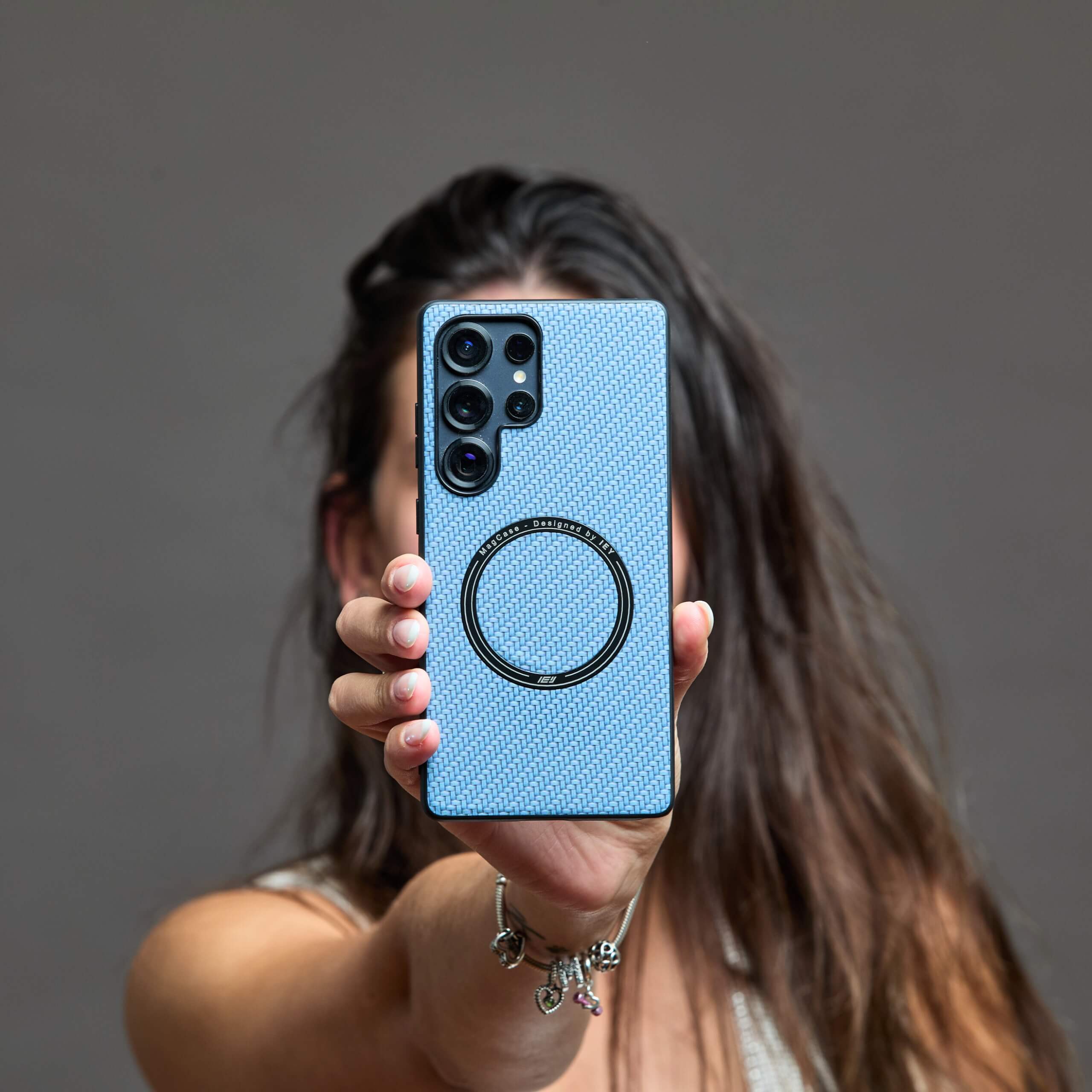 Mujer sosteniendo un Samsung con funda MagSafe azul frente a su rostro, fondo beige claro, enfoque en el producto #color_azul