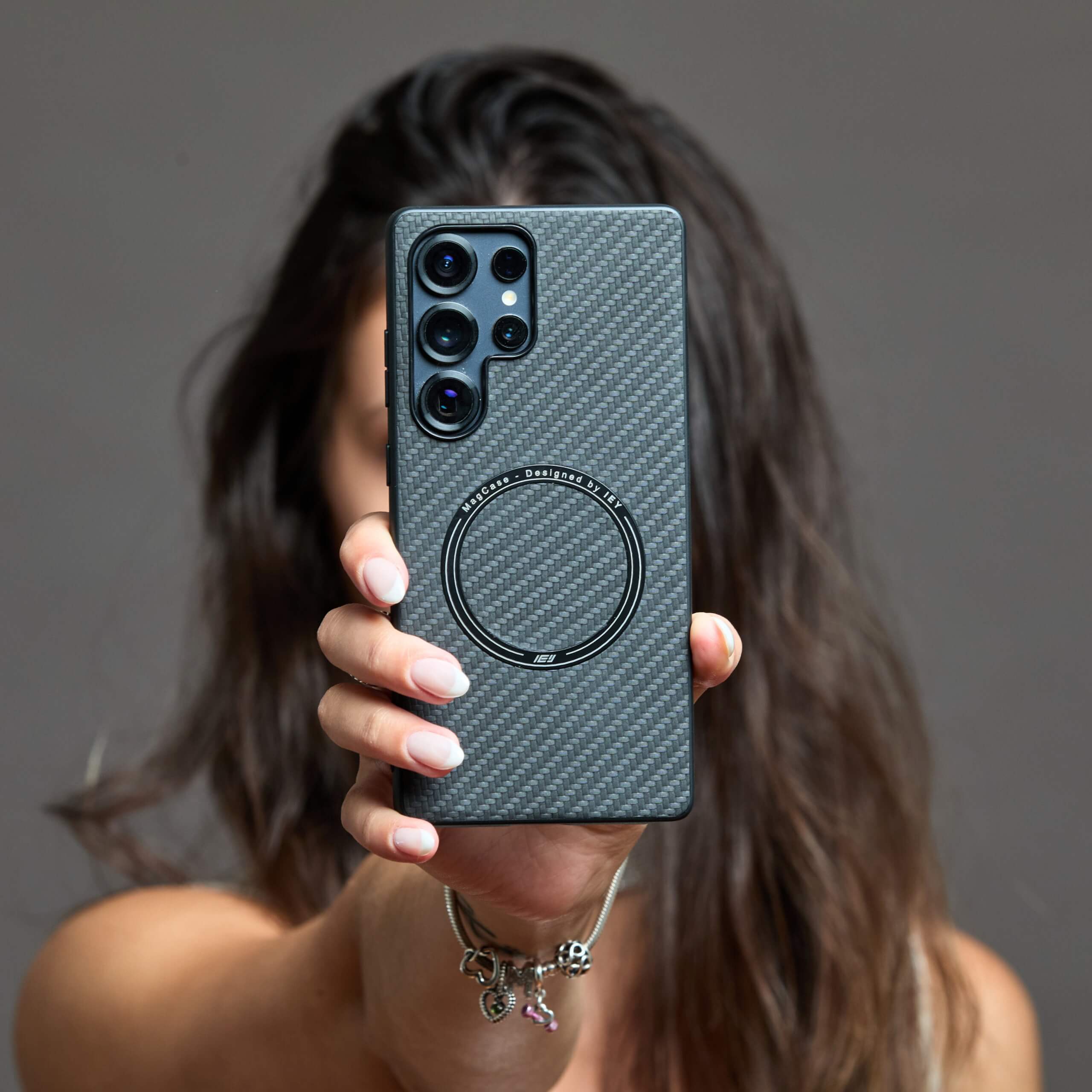 Mujer con suéter blanco sosteniendo un Samsung con funda MagSafe de fibra de carbono frente a la cámara, en gesto de selfie #color_negro