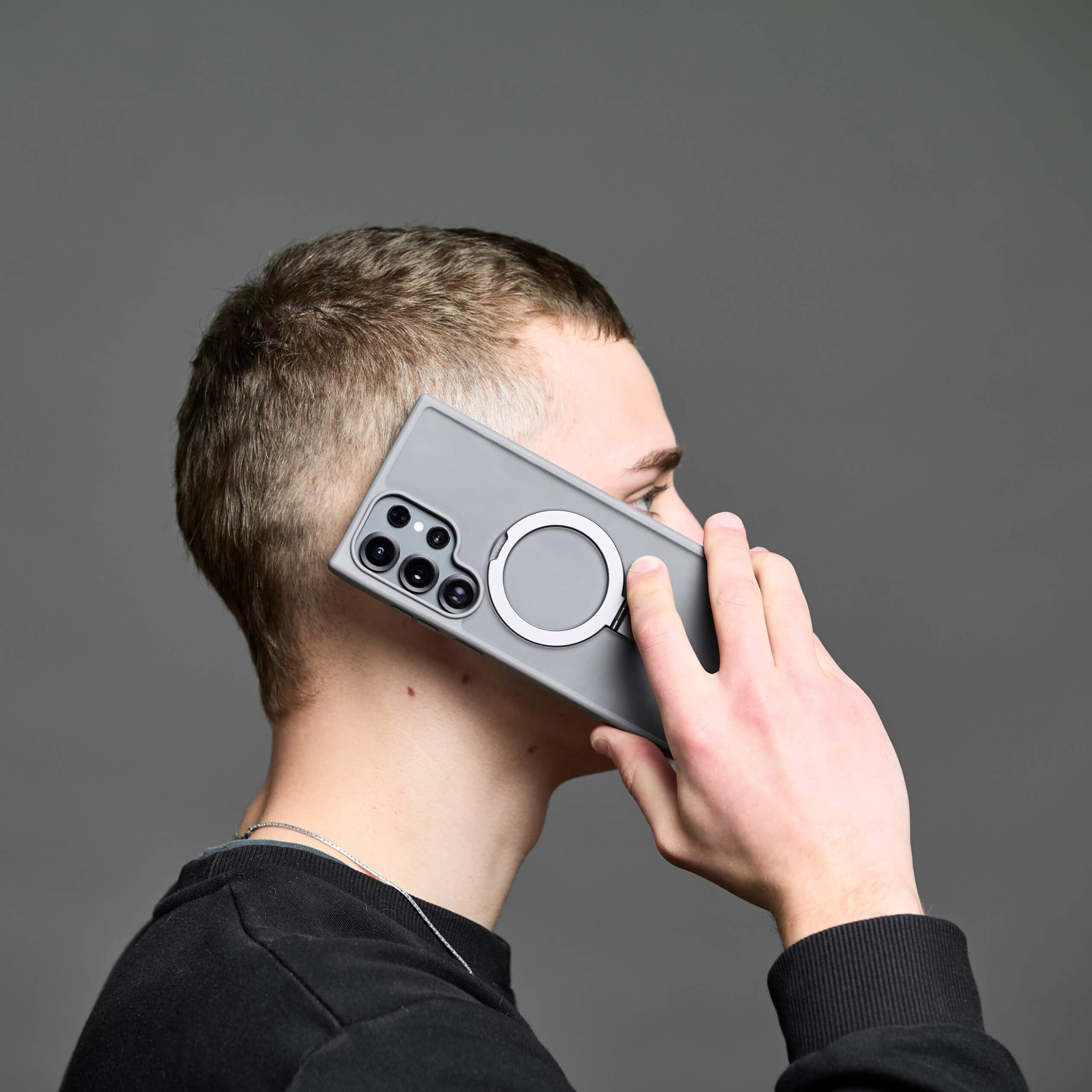Hombre mostrando el teléfono en la oreja con funda y aro magnético, fondo gris.