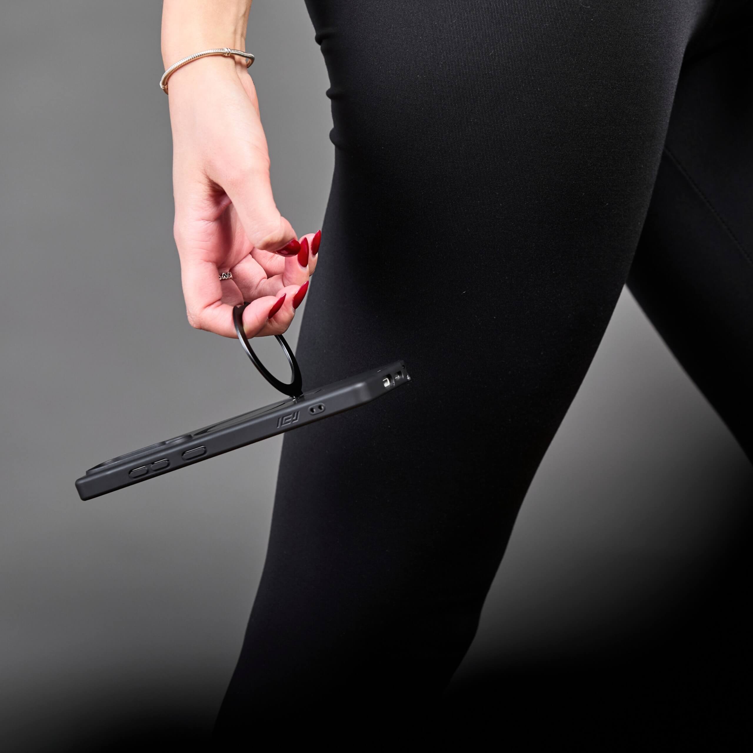 Persona sosteniendo el teléfono con funda y aro magnético desde el aro, al costado de la pierna. #color_negro