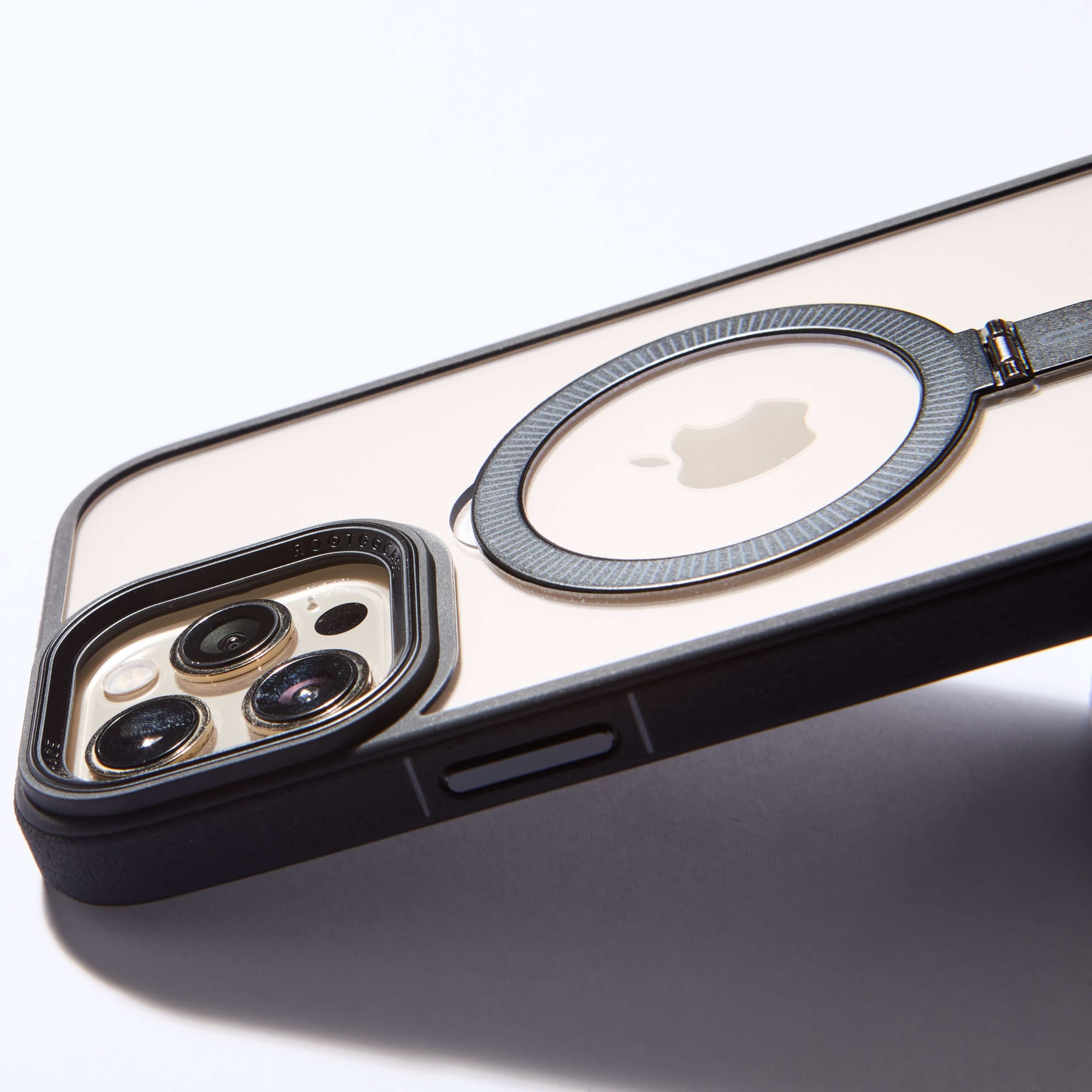 Funda MagSafe negra con anillo desplegable, vista en detalle sobre fondo blanco mostrando cámara y logo Apple. #color_negro