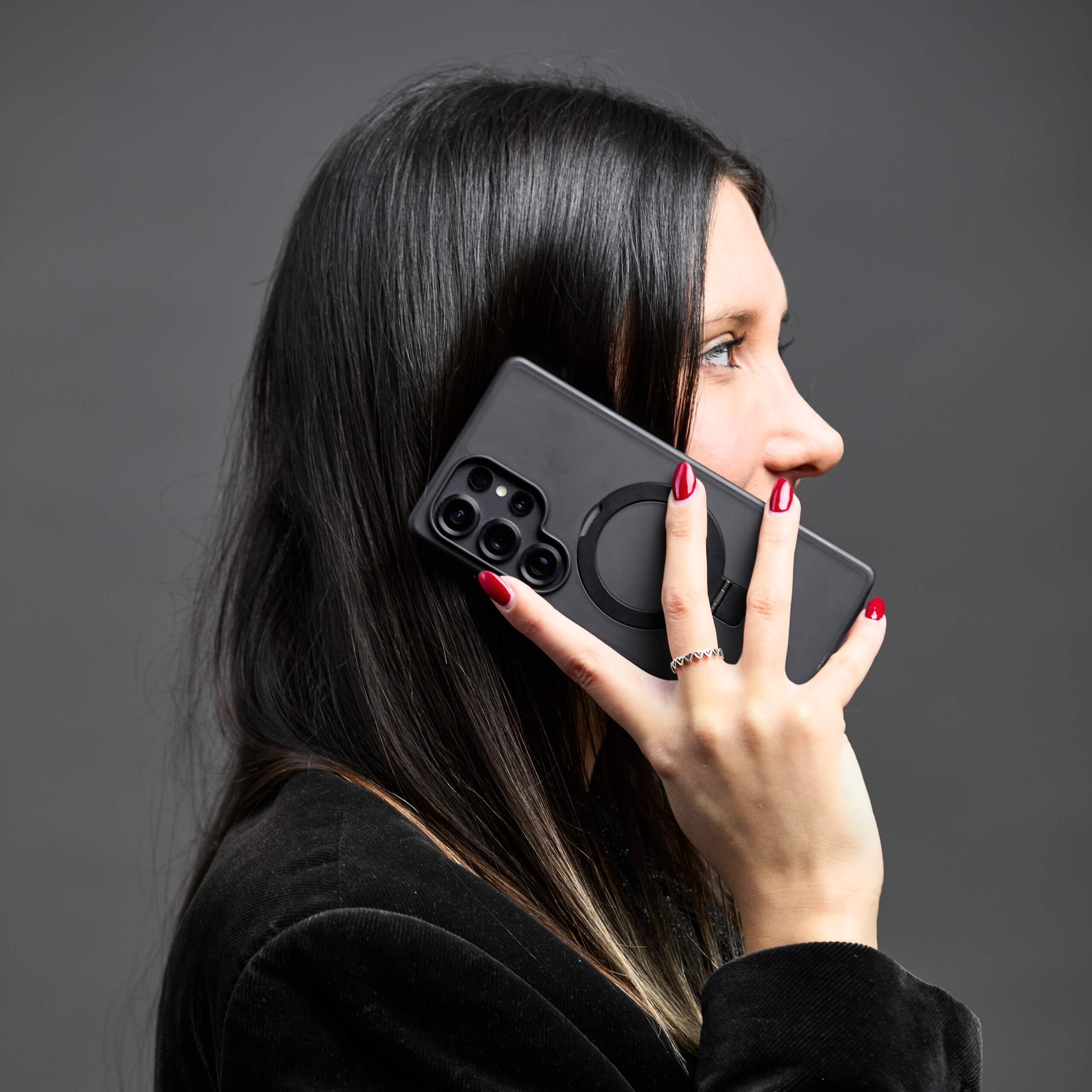 Mujer hablando por teléfono Samsung S25 Ultra con funda con aro magnético, fondo gris oscuro. #color_negro