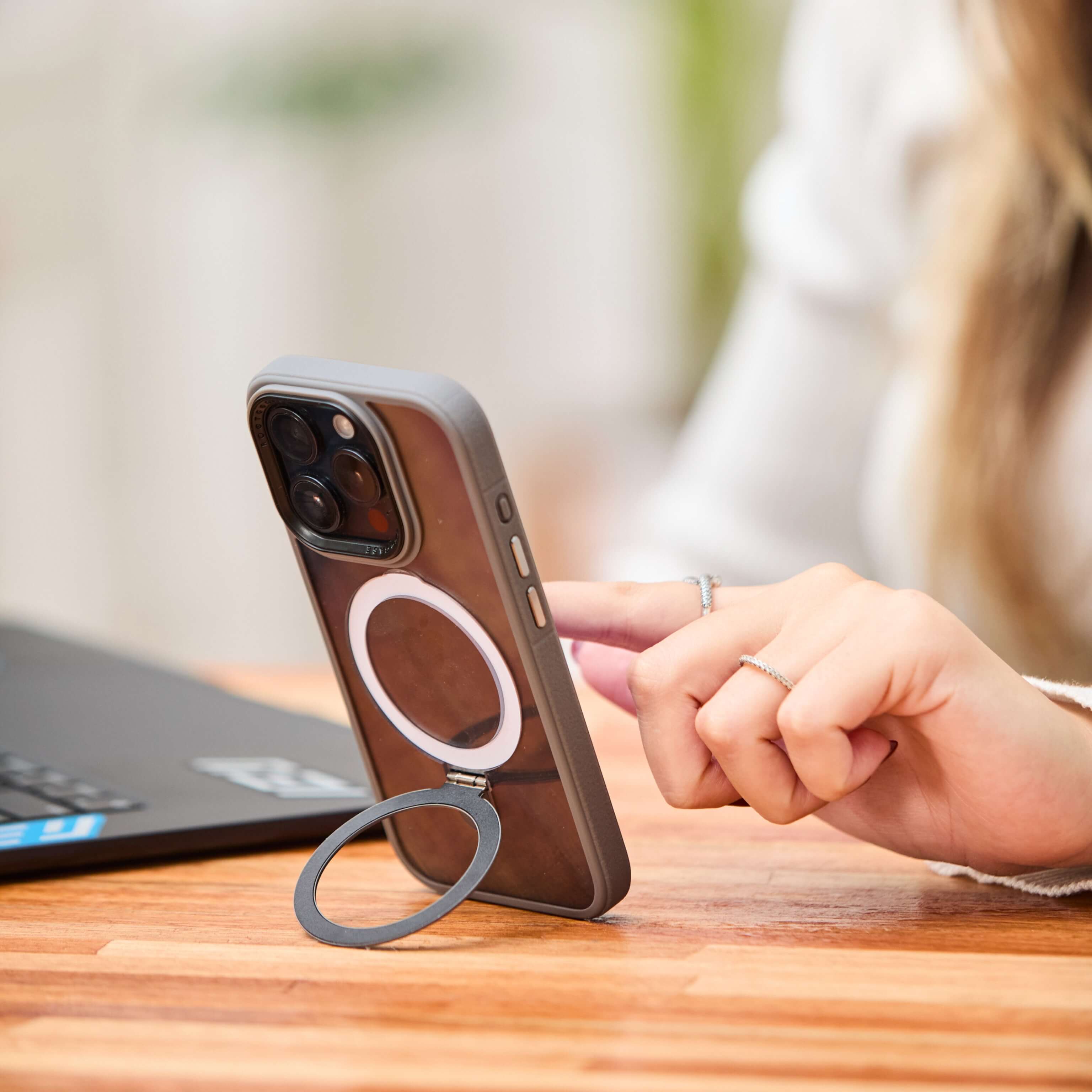 Funda gris con anillo trasero desplegado sosteniendo el teléfono en posición vertical mientras una mujer lo utiliza sobre un escritorio. #color_gris