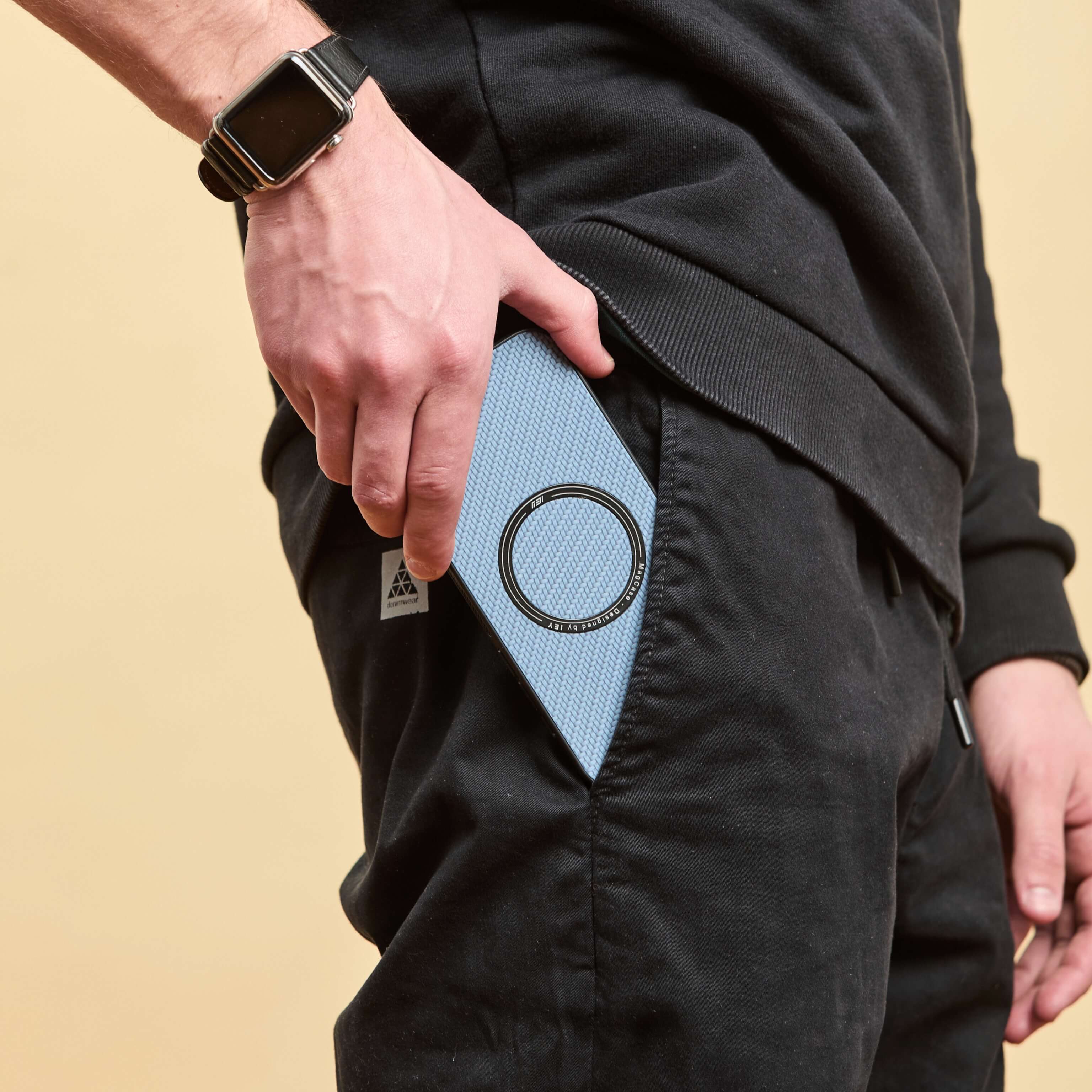 Mano masculina sacando un iPhone con funda MagSafe azul del bolsillo lateral de un pantalón negro, fondo beige claro #color_azul