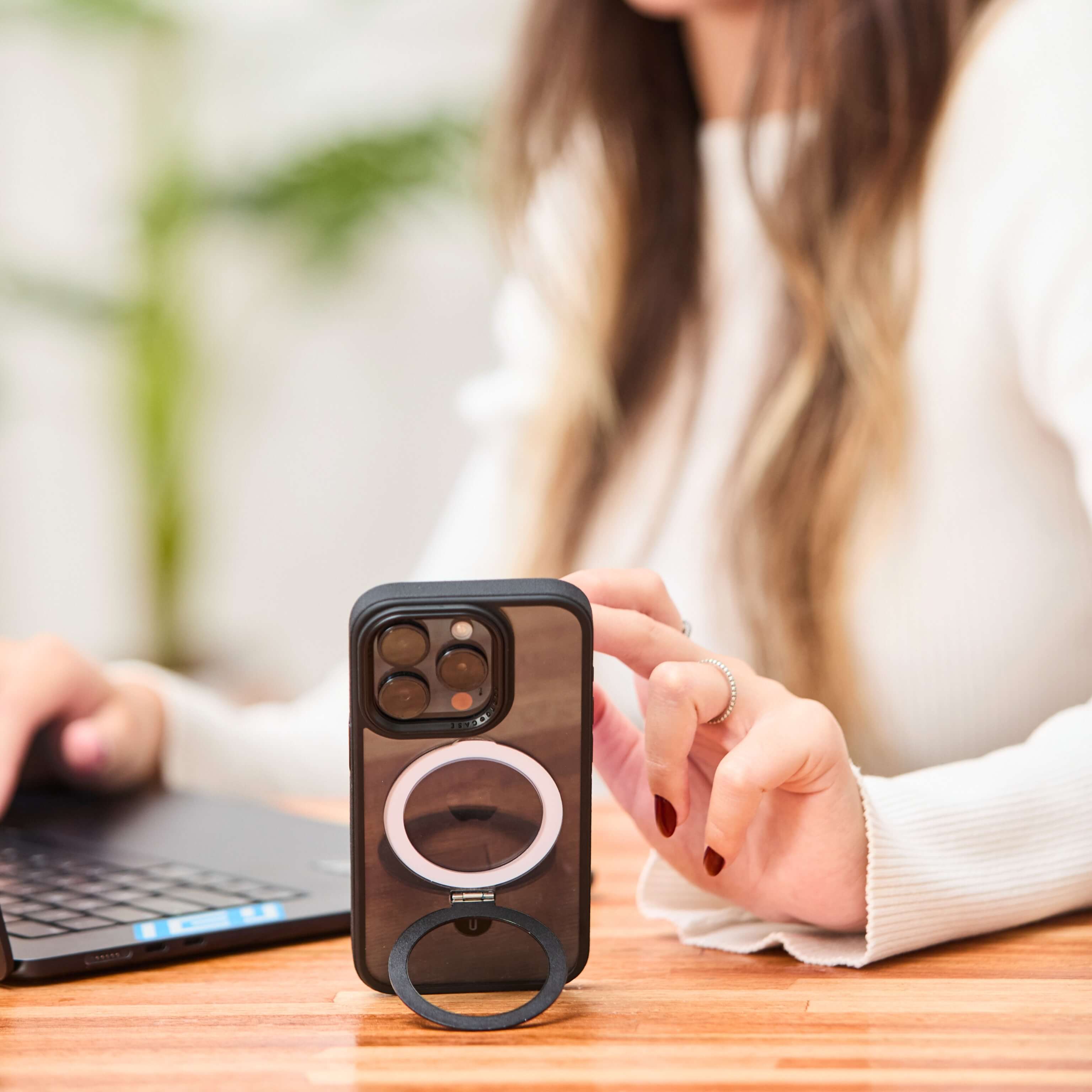Funda negra con anillo desplegado sosteniendo el teléfono en posición vertical sobre escritorio mientras una mujer lo utiliza. #color_negro
