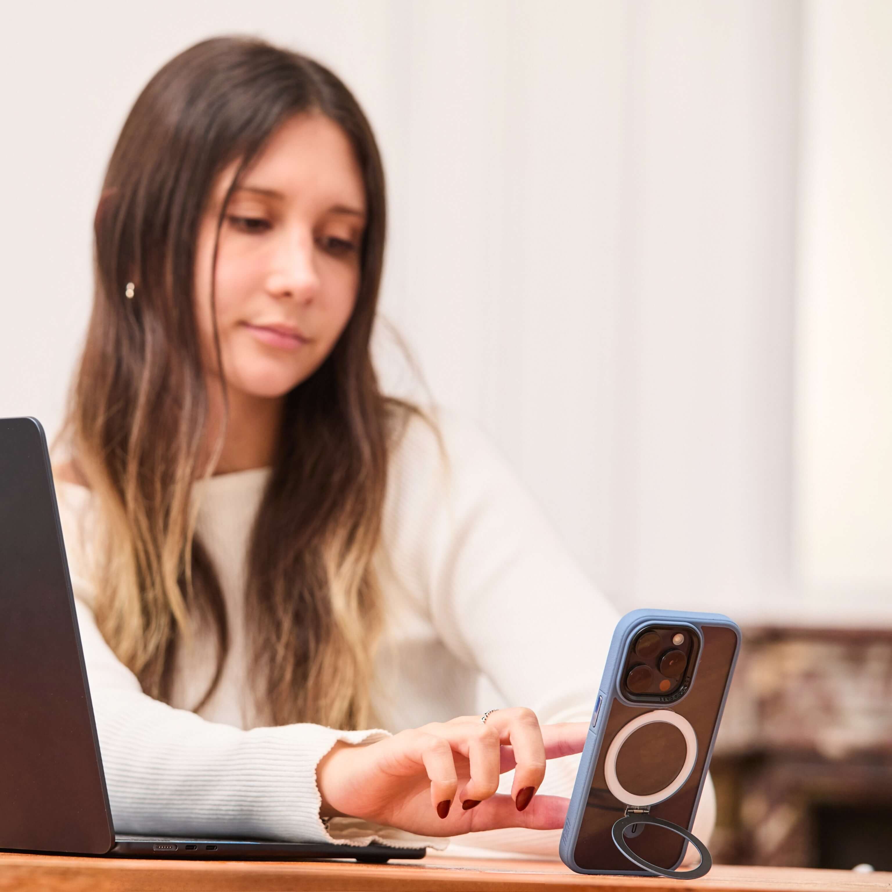 Mujer interactúa con un iPhone azul sostenido por su anillo soporte, apoyado verticalmente sobre escritorio junto a una notebook. #color_azul