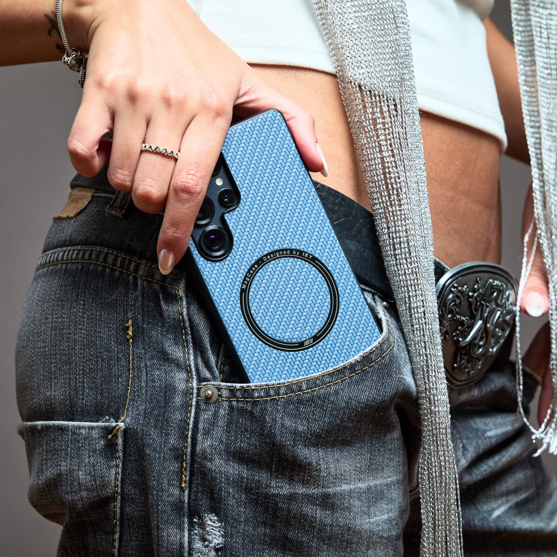 Mano femenina sacando un Samsung con funda MagSafe azul del bolsillo lateral de un pantalón negro, fondo beige claro #color_azul
