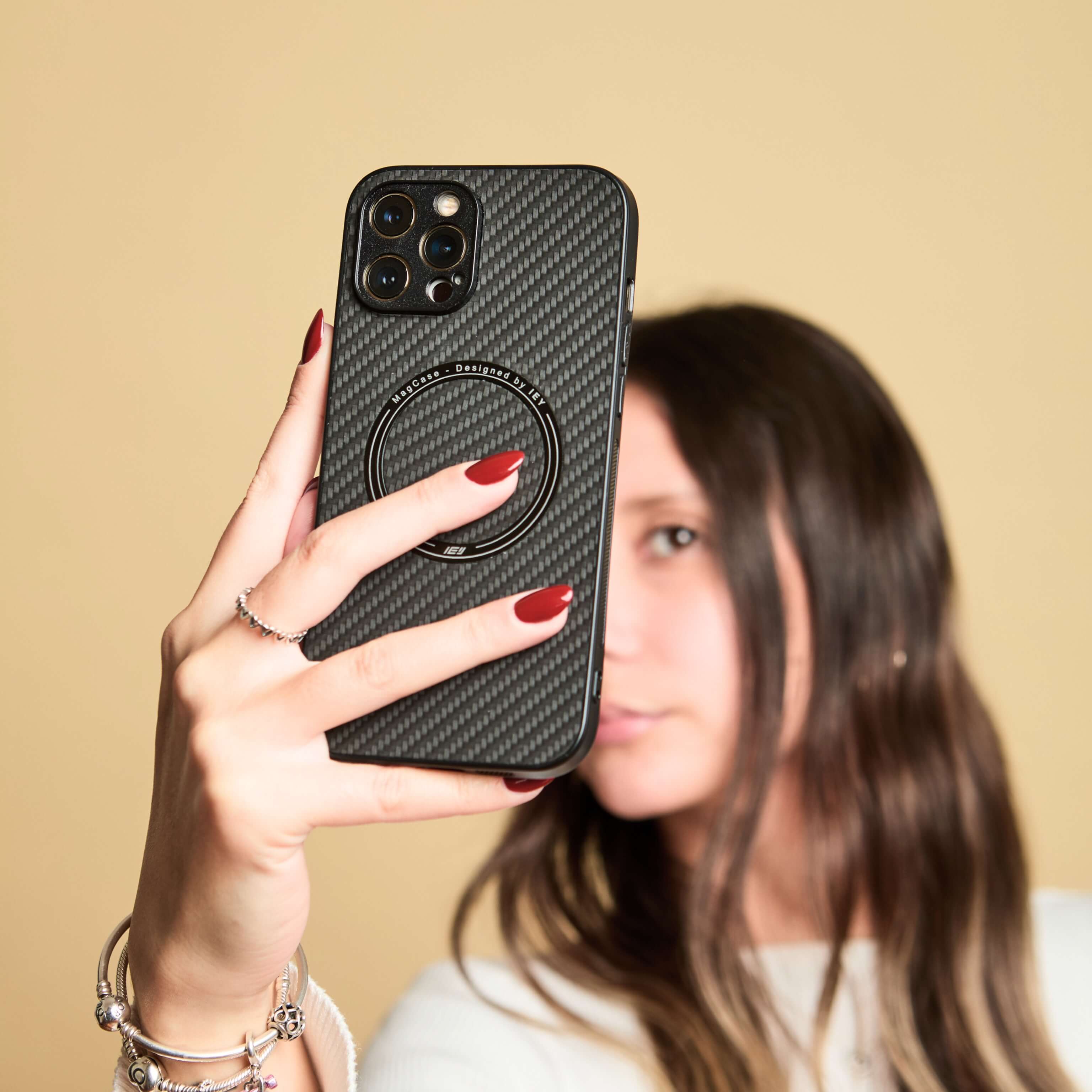 Mujer sosteniendo su teléfono frente al rostro con funda MagSafe negra de textura carbono, sobre fondo beige #color_negro