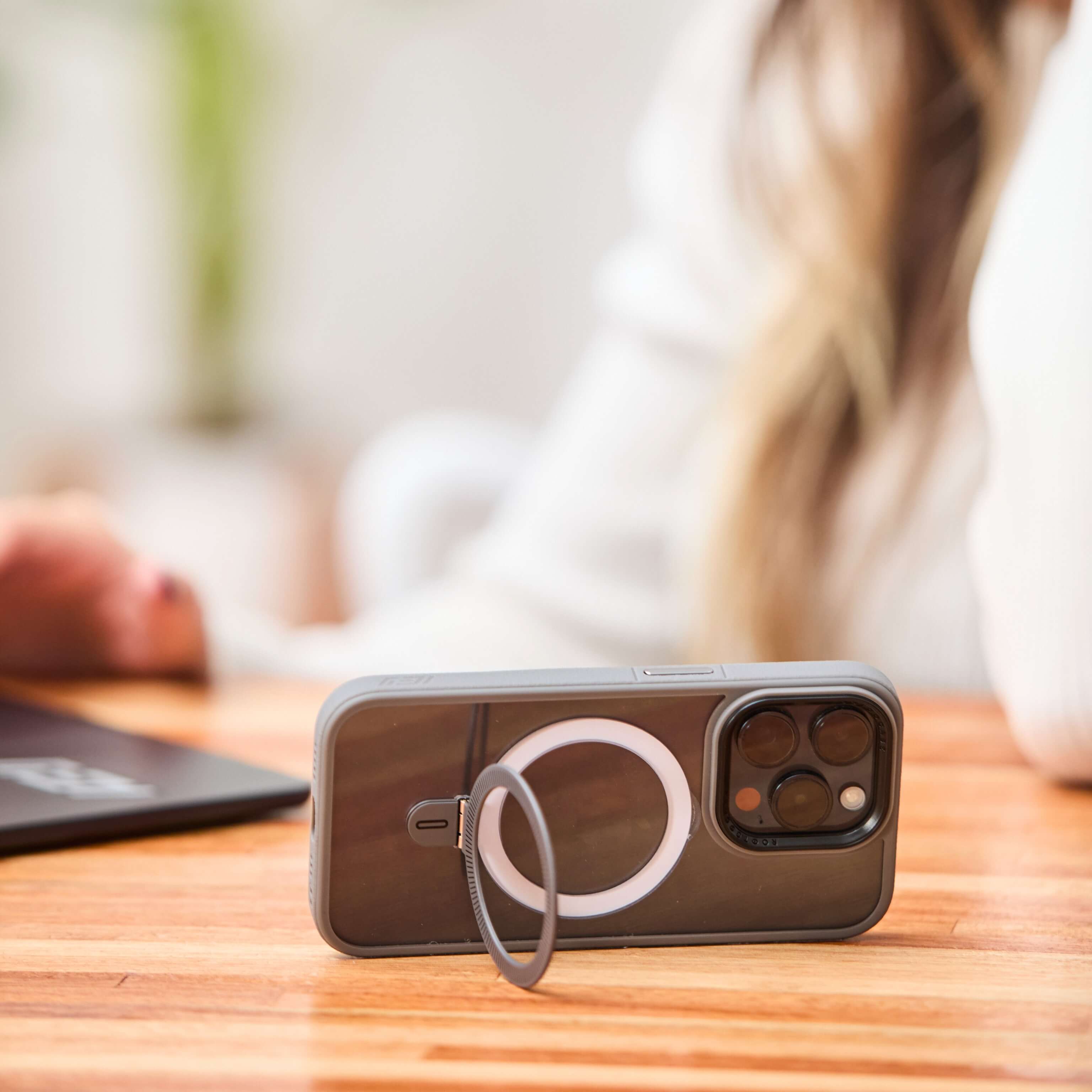 Funda gris con anillo trasero desplegado sosteniendo el teléfono en posición horizontal sobre una mesa, con una mujer desenfocada al fondo. #color_gris