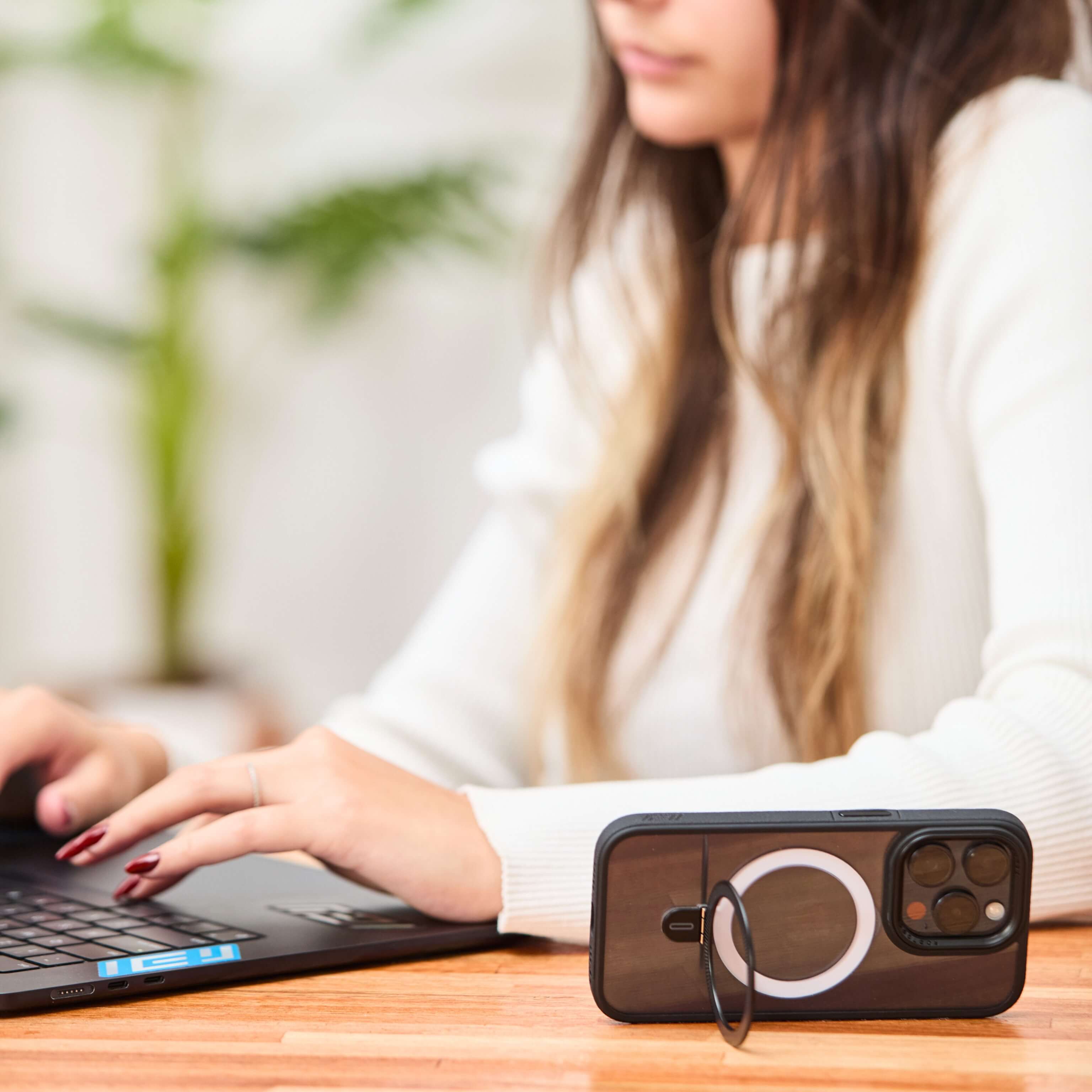 Mujer trabajando en laptop con iPhone apoyado horizontalmente usando el anillo trasero como soporte, sobre escritorio de madera. #color_negro