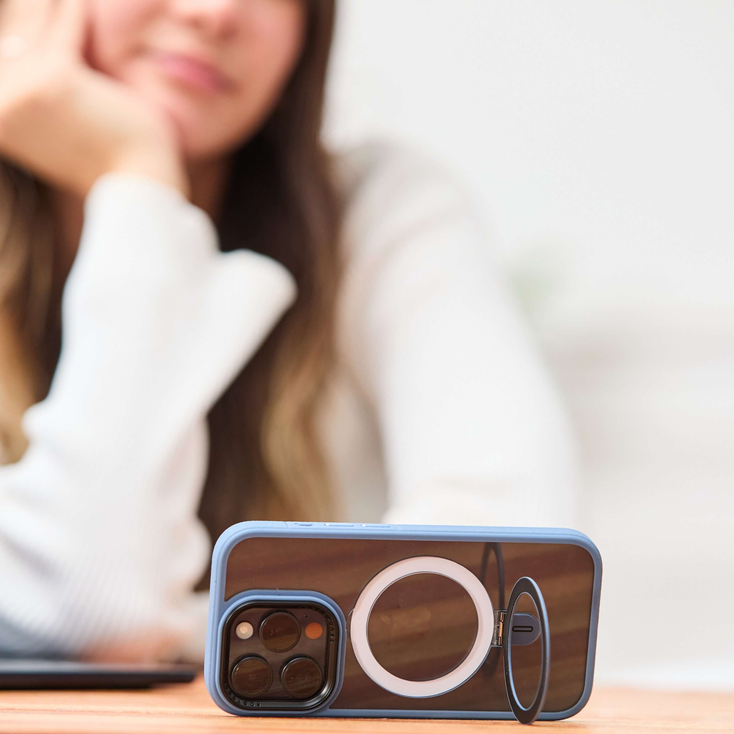 Mujer de fondo desenfocado observa un iPhone apoyado horizontalmente con anillo desplegado, sobre escritorio de madera. #color_azul