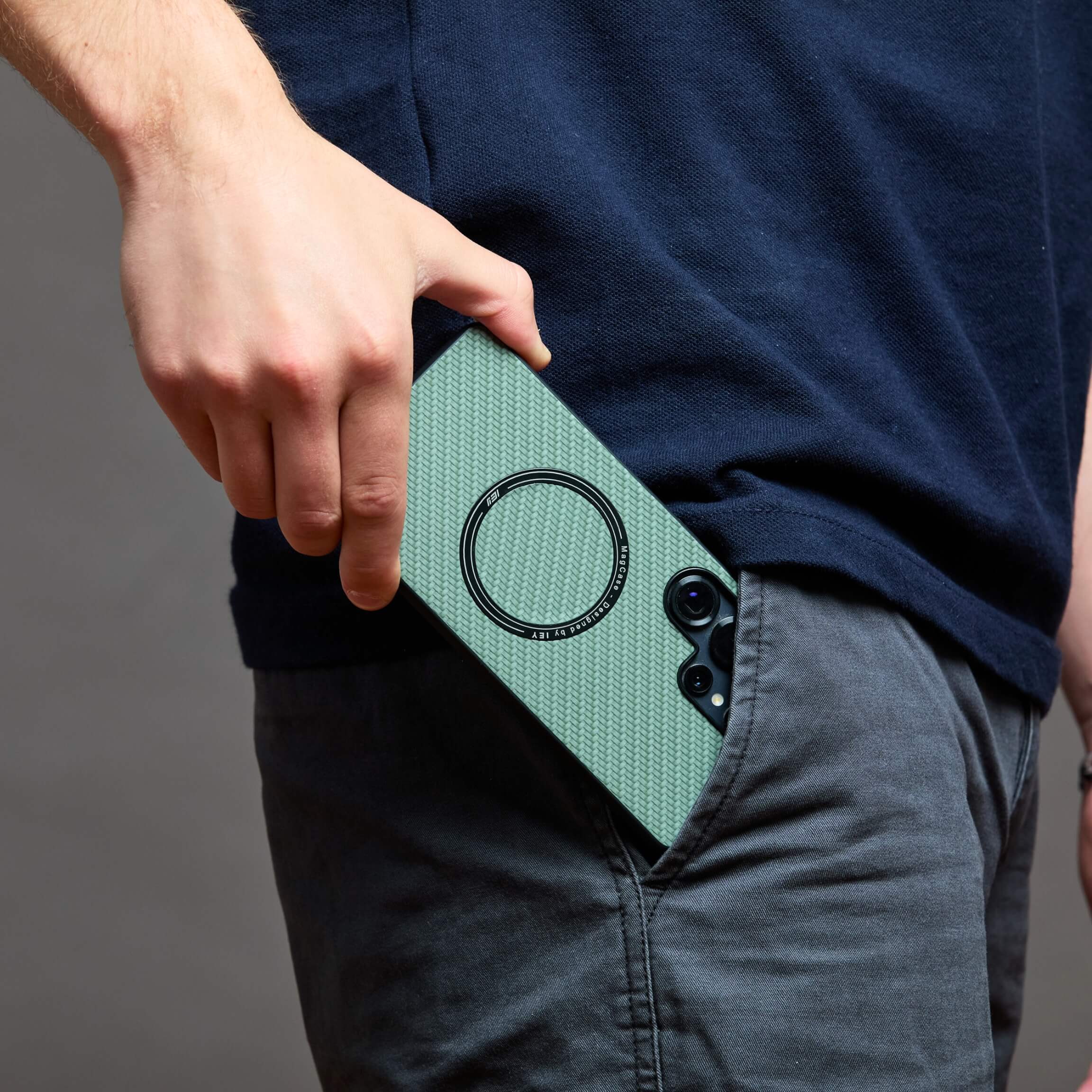 Funda MagSafe Samsung verde con textura de carbono, sostenida en la mano de un hombre con pantalón negro y reloj, en plano detalle lateral #color_verde