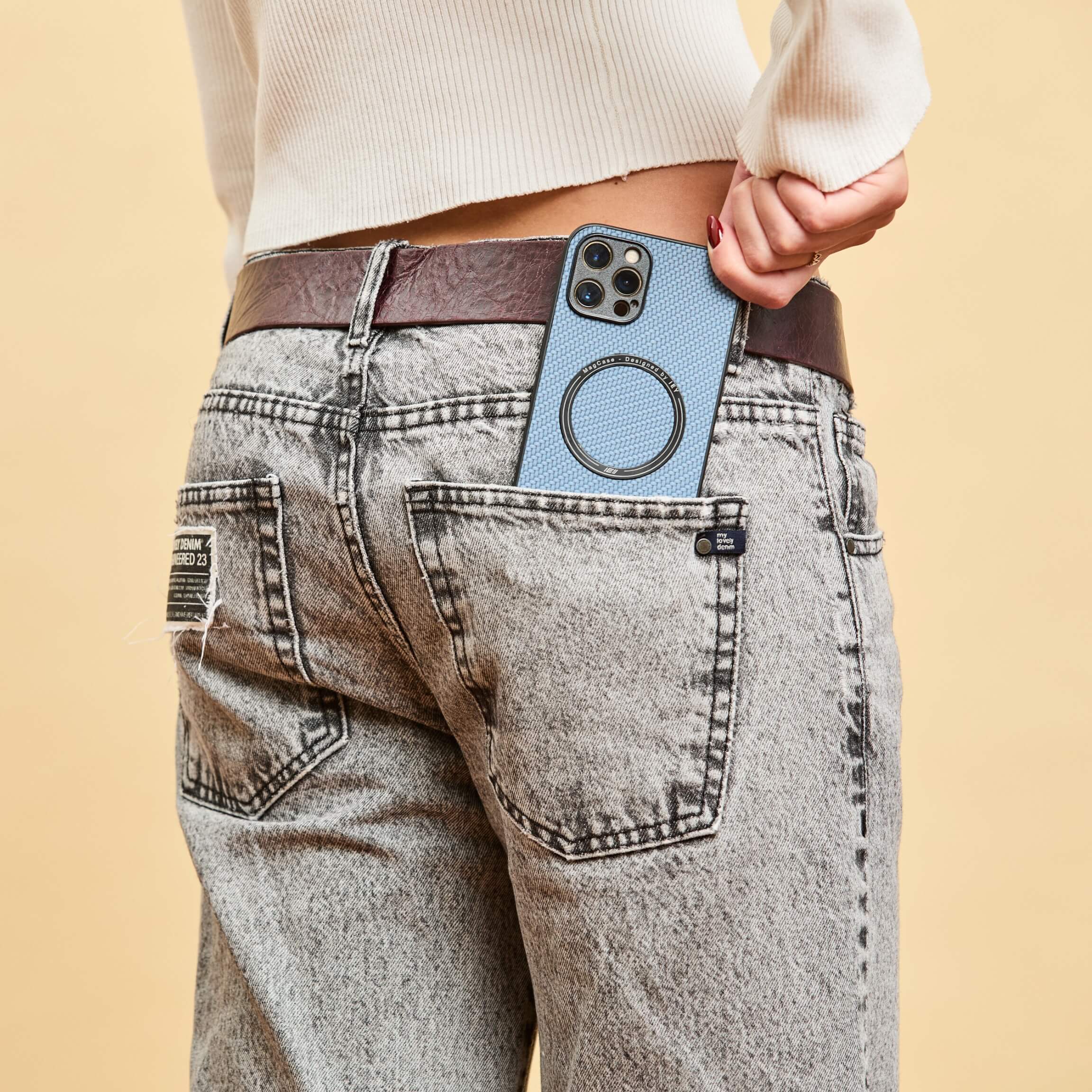 Funda MagSafe azul en iPhone, asomando desde el bolsillo trasero de un jean gris claro, con cinturón burdeos y suéter blanco #color_azul