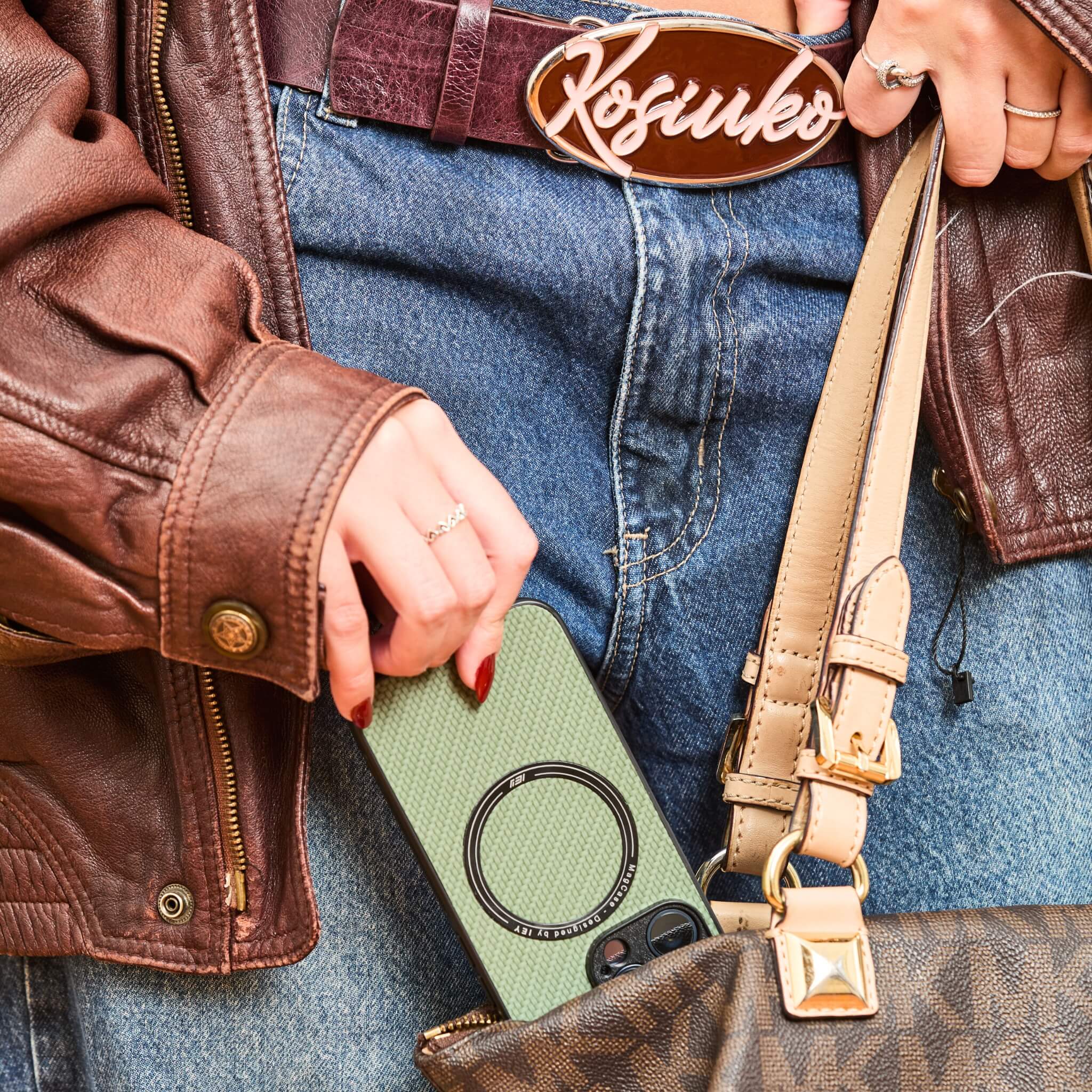 Funda MagSafe verde con textura símil carbono, en mano femenina colocándola dentro de una cartera, estilo urbano con campera de cuero y jeans #color_verde