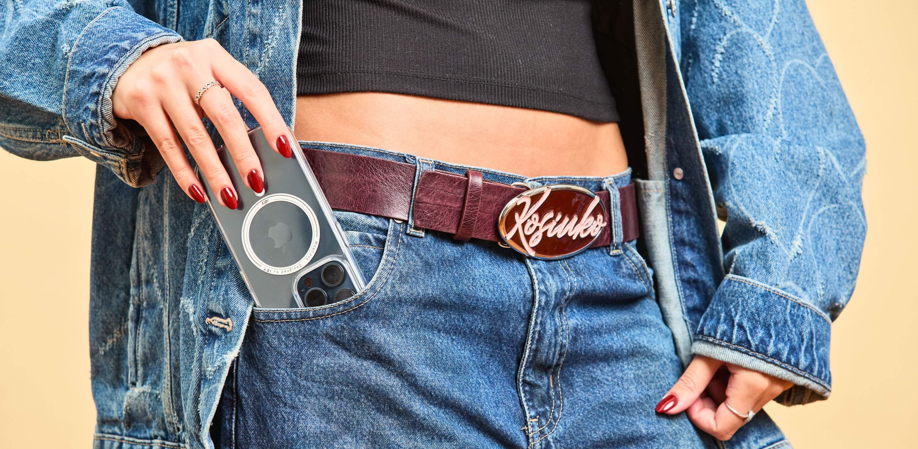Mujer guardando un iPhone con funda transparente MagSafe en el bolsillo delantero de un jean, con cinturón burdeos y look total denim