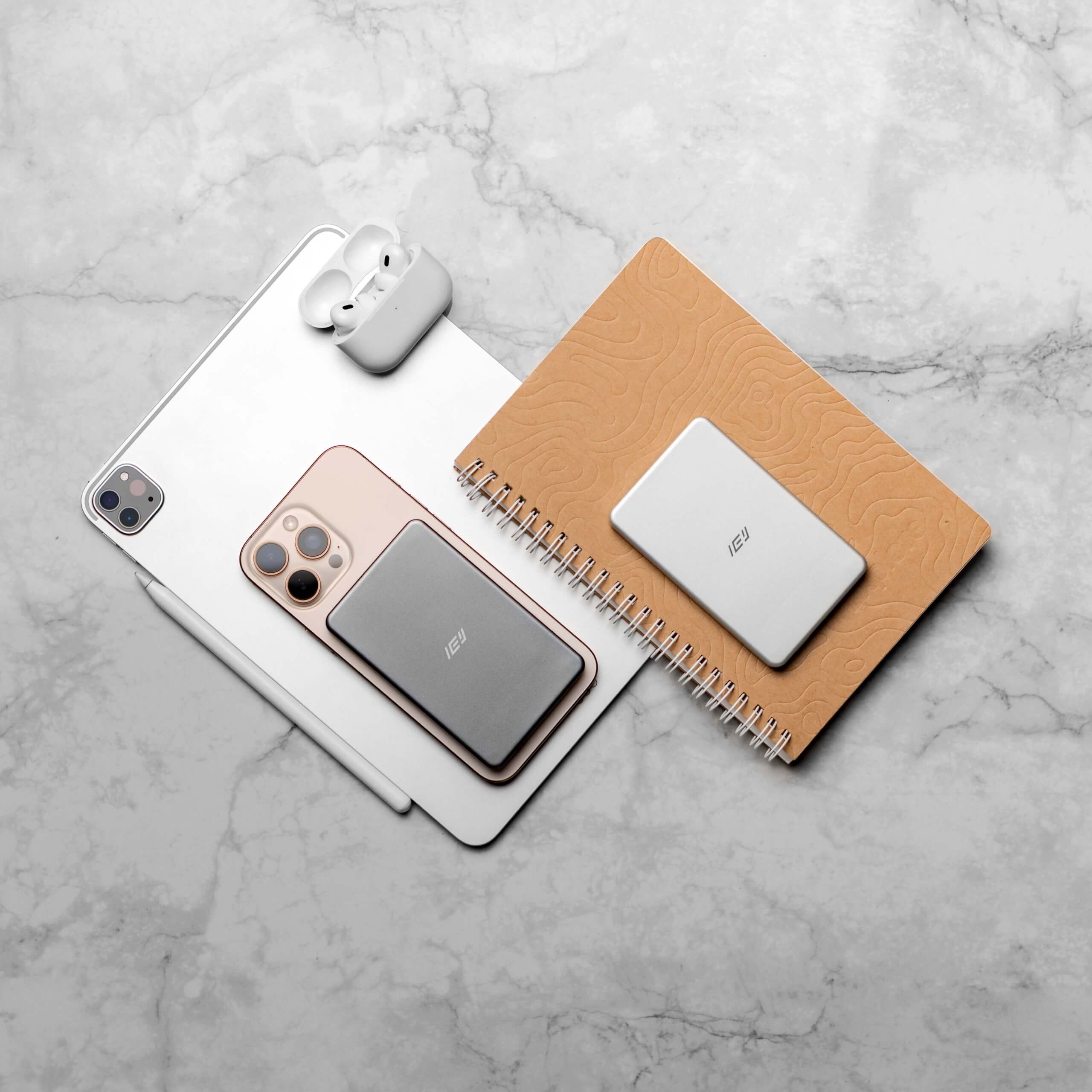 Batería inalámbrica MagSafe IEY ultrafina en color gris y blanco, junto a iPhone, iPad y AirPods, estilo lifestyle sobre mesa de mármol.
