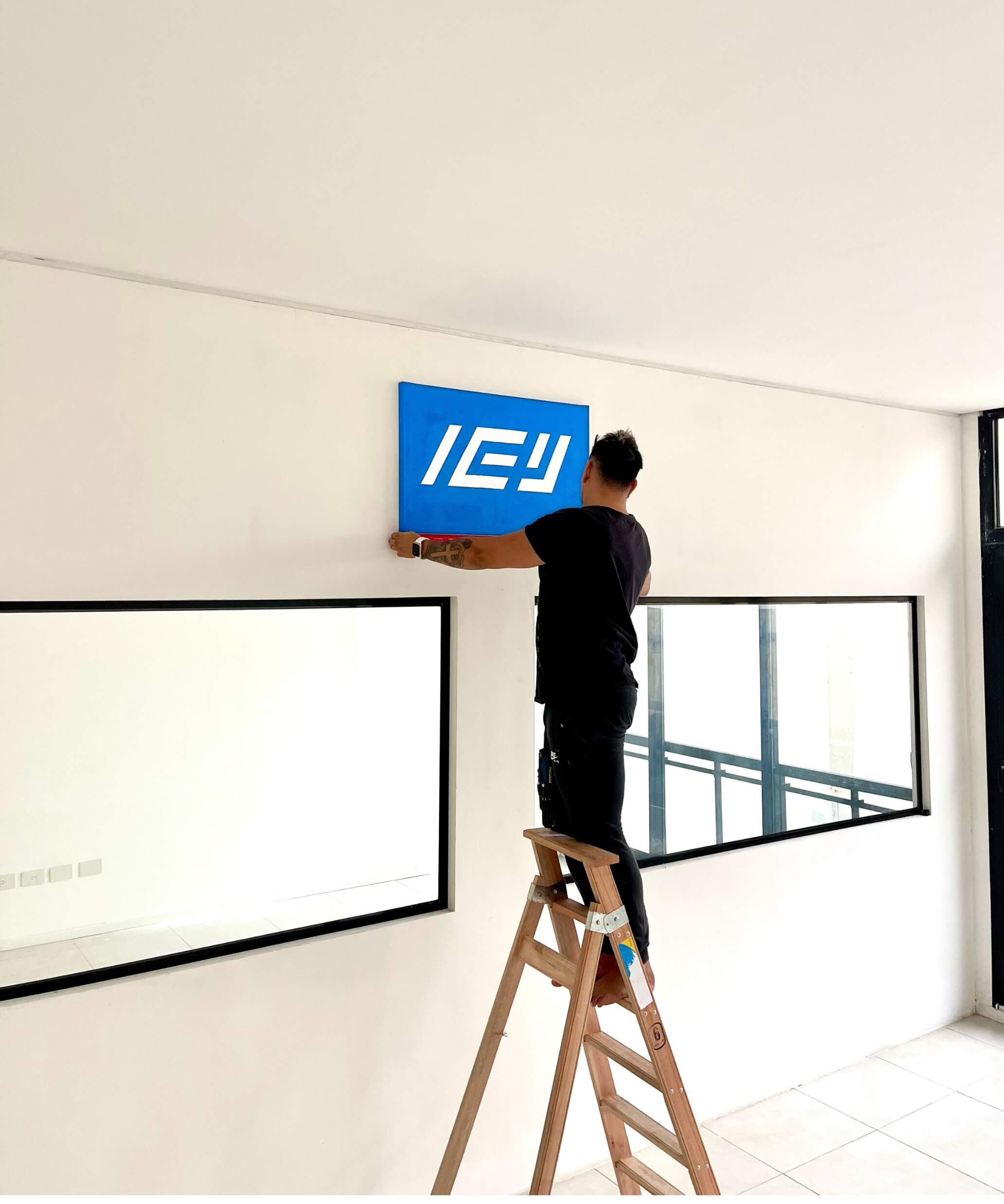Persona instalando cartel con logotipo de IEY sobre pared blanca en oficina moderna, representando identidad de marca