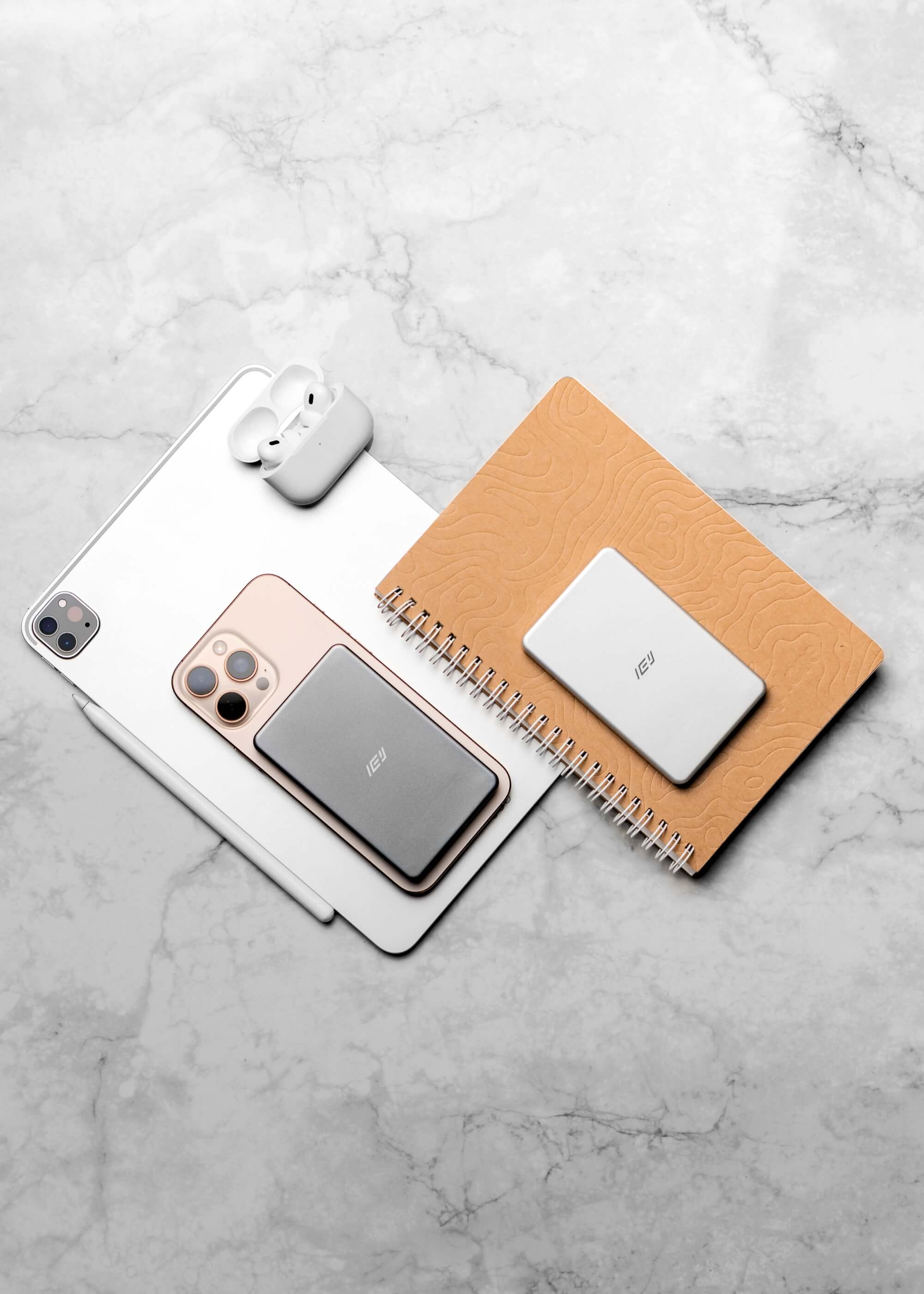 Batería inalámbrica MagSafe IEY ultrafina en color gris y blanco, junto a iPhone, iPad y AirPods, estilo lifestyle sobre mesa de mármol.