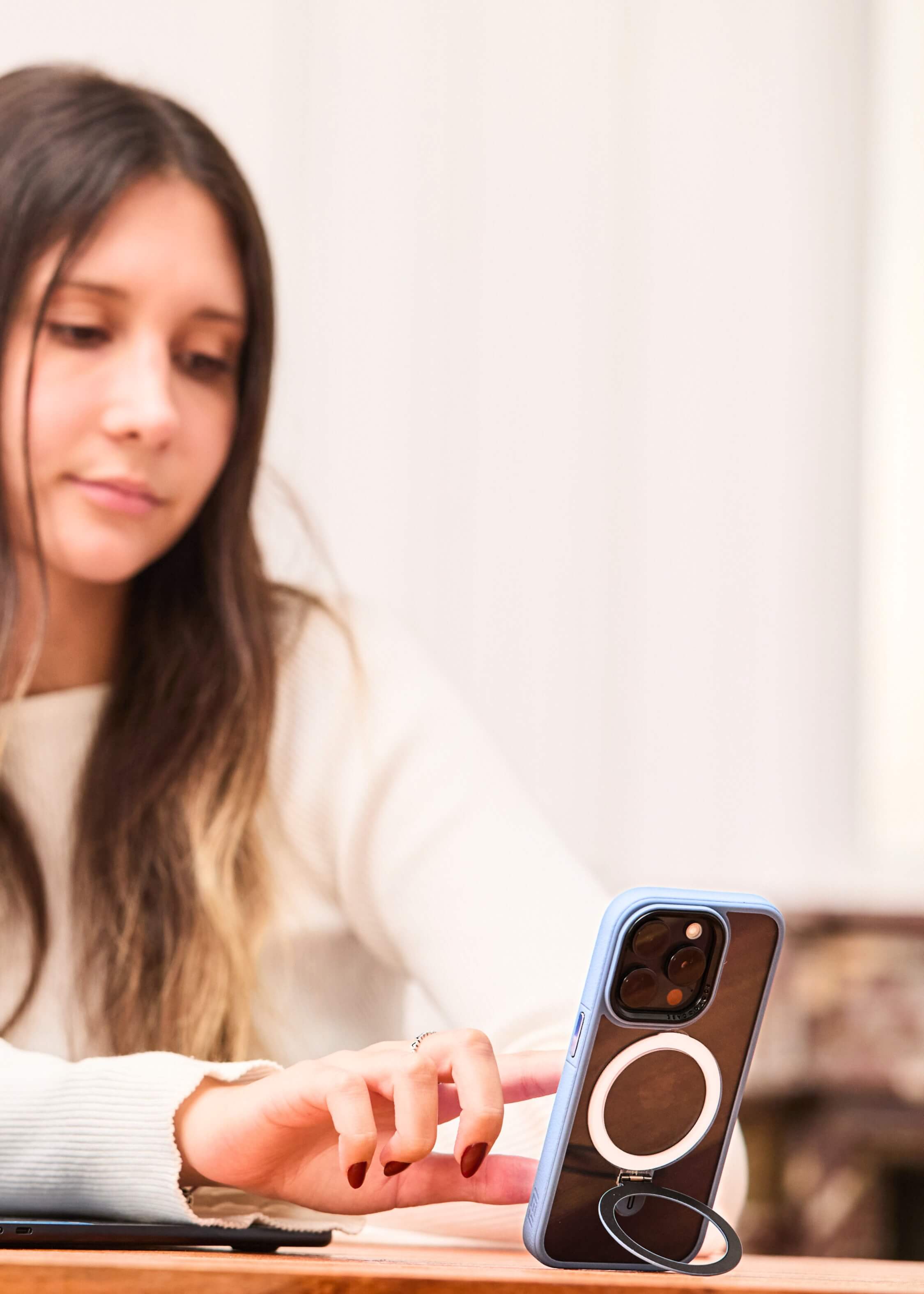 Mujer interactúa con un iPhone con Funda Azul sostenido por su anillo soporte, apoyado verticalmente sobre escritorio junto a una notebook.