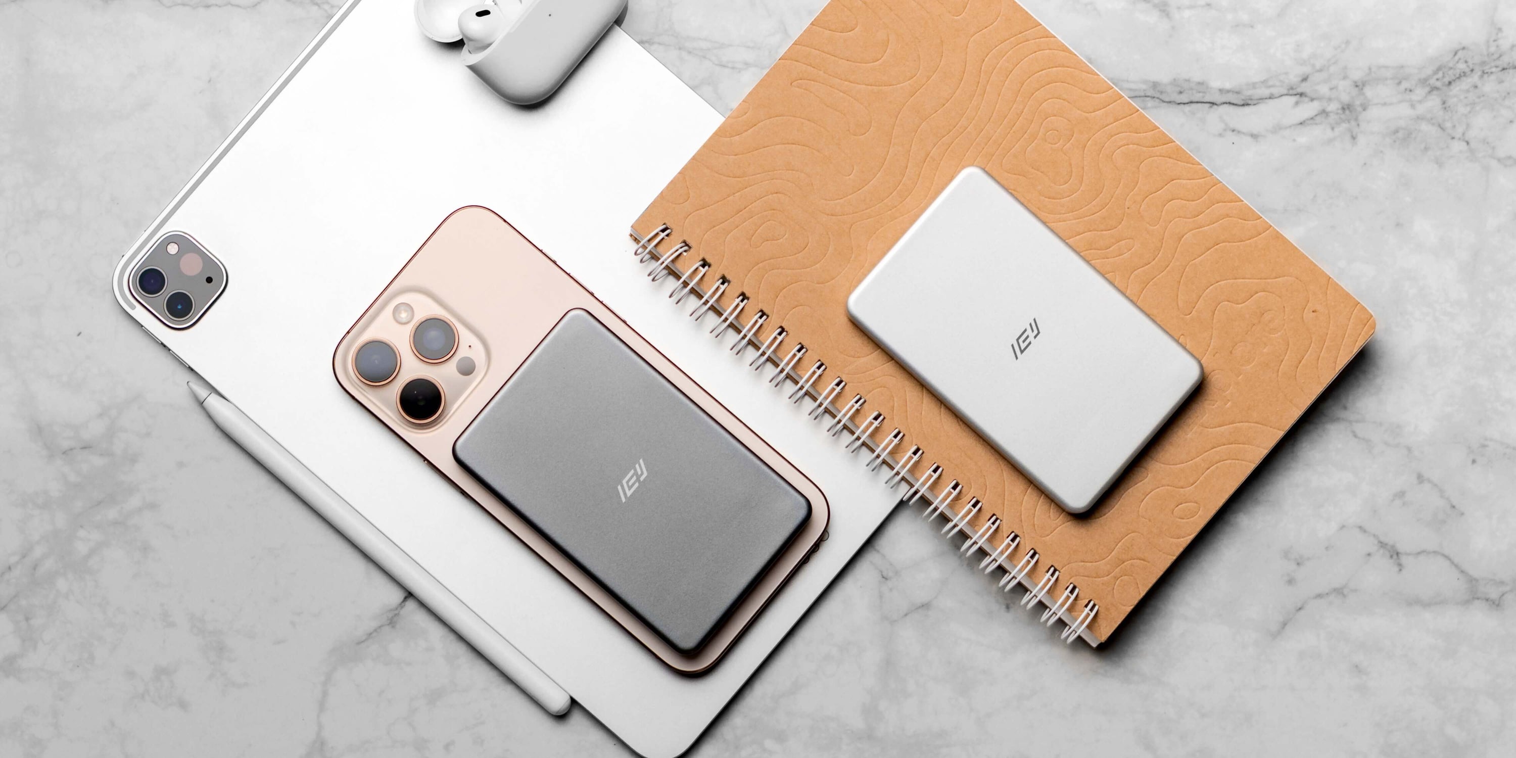 Batería inalámbrica MagSafe IEY ultrafina en color gris y blanco, junto a iPhone, iPad y AirPods, estilo lifestyle sobre mesa de mármol.