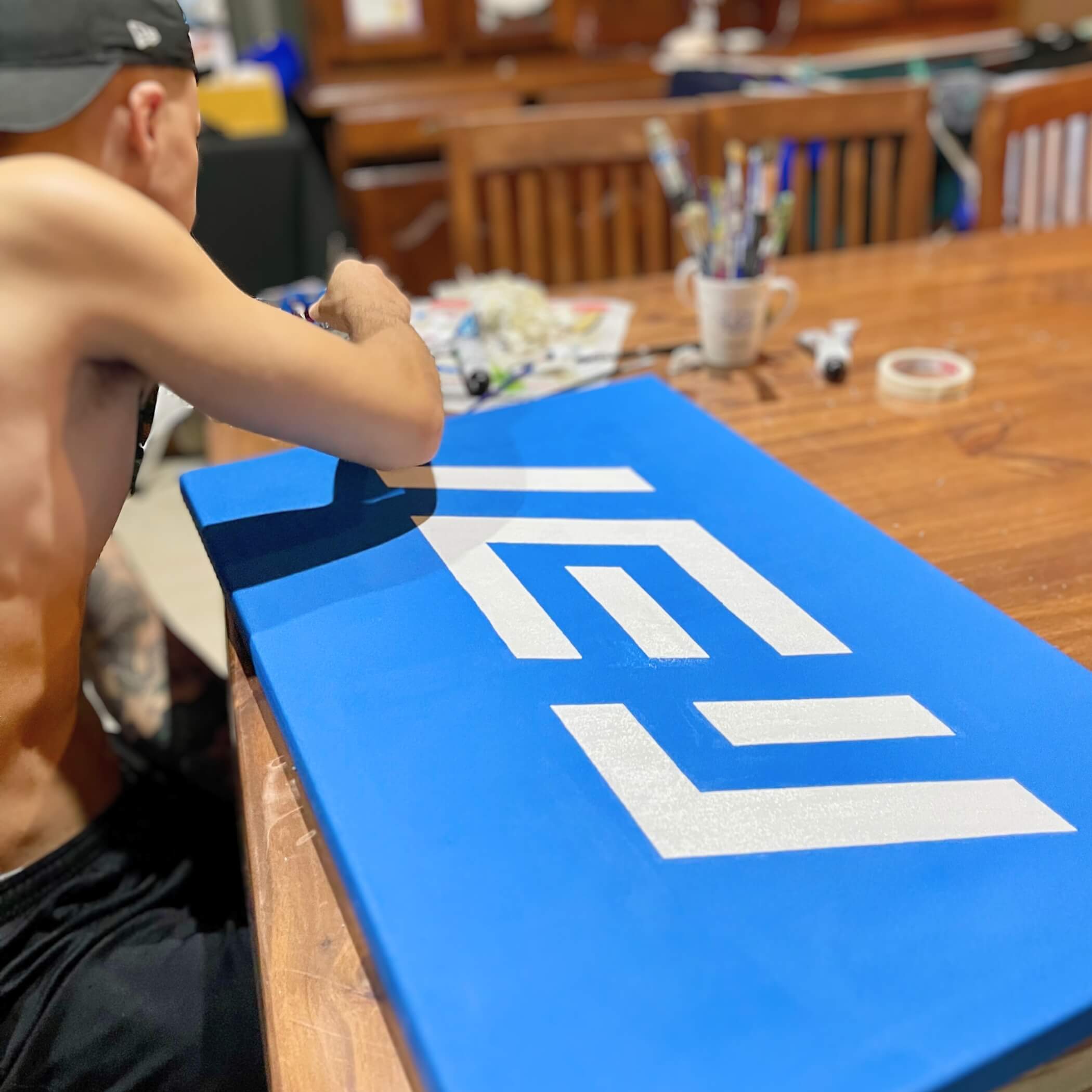 Persona pintando a mano el logotipo de IEY sobre lienzo azul, mostrando el proceso creativo y artesanal detrás de la identidad visual de la marca